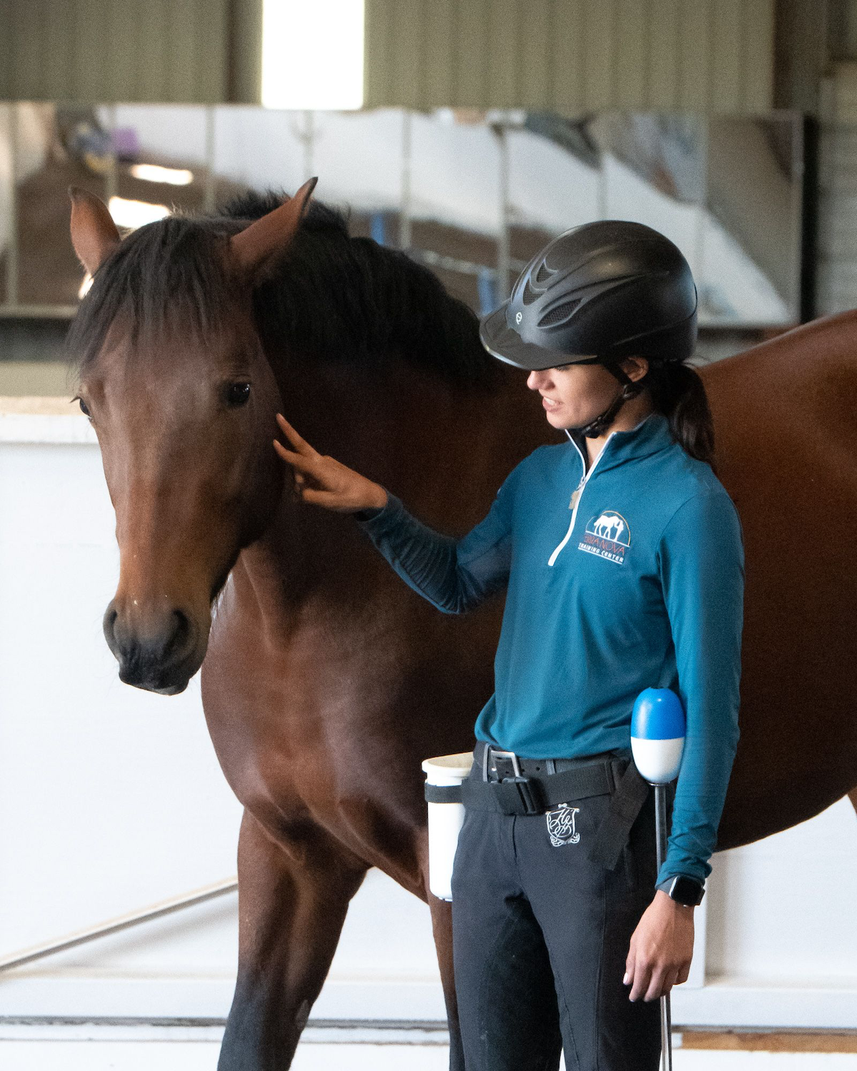 Jasmine touches the Taz, a bay Mustang's cheek with two fingers. She wears a riding helmet and teal shirt in an indoor arena, demonstrating a clear, quiet connection focused while training.